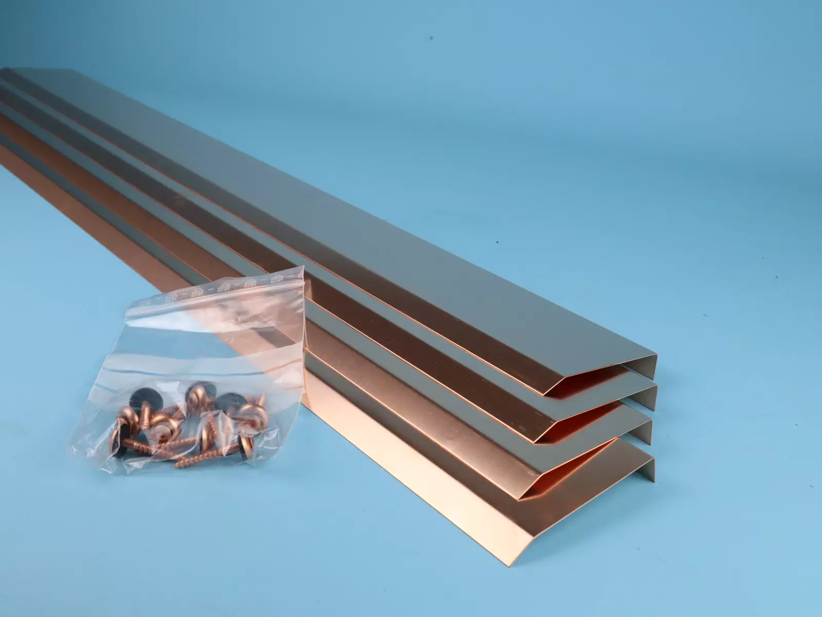 Vier, längliche, kupferfarbene Metallprofile mit einem nach unten gebogenen Ende liegen übereinander. Neben ihnen befindet sich eine Plastikbeutel mit Schrauben und Unterlegscheiben. Der Hintergrund ist ein einfarbiges, hellblaues Bild.