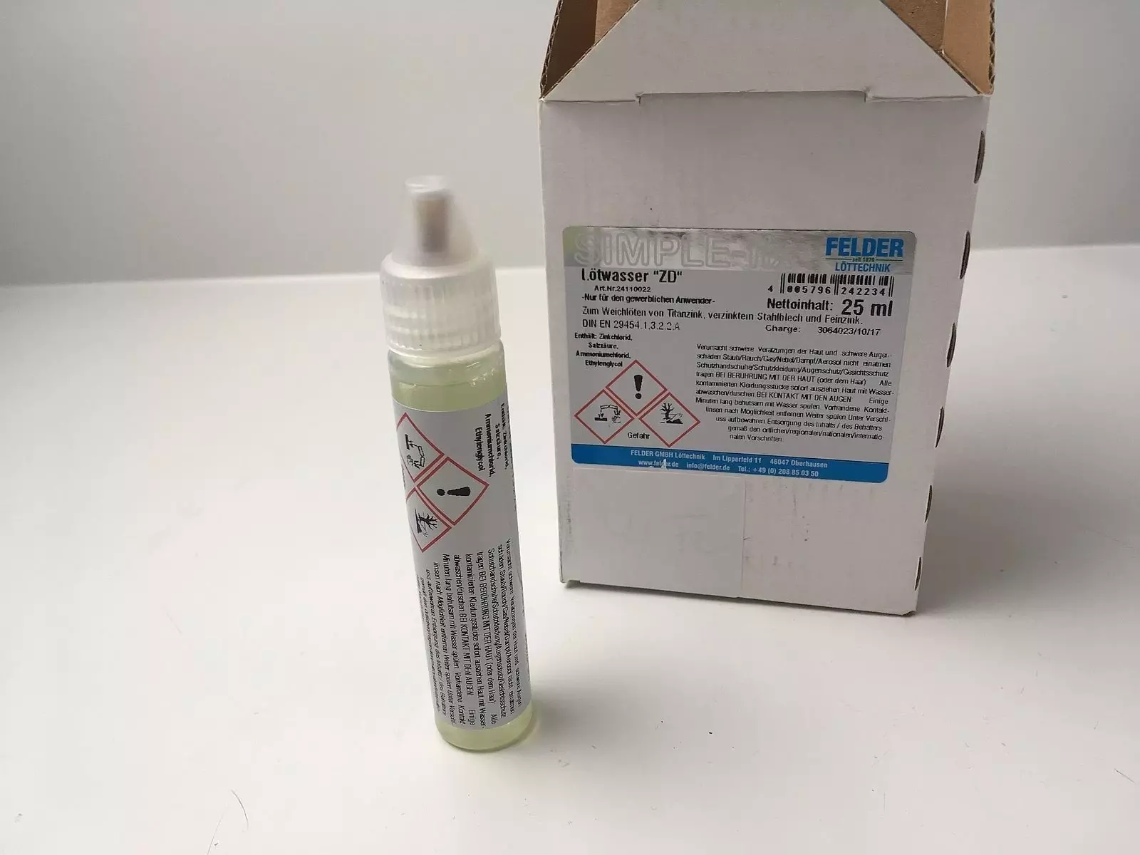 Eine Flasche mit Lötdrahtflussmittel steht neben einer weißen Verpackung. Die Flasche hat eine grüne Flüssigkeit, eine spitz zulaufende Kappe und Sicherheitswarnungen auf dem Etikett. Die Verpackung zeigt Informationen über das Produkt und dessen Anwendun
