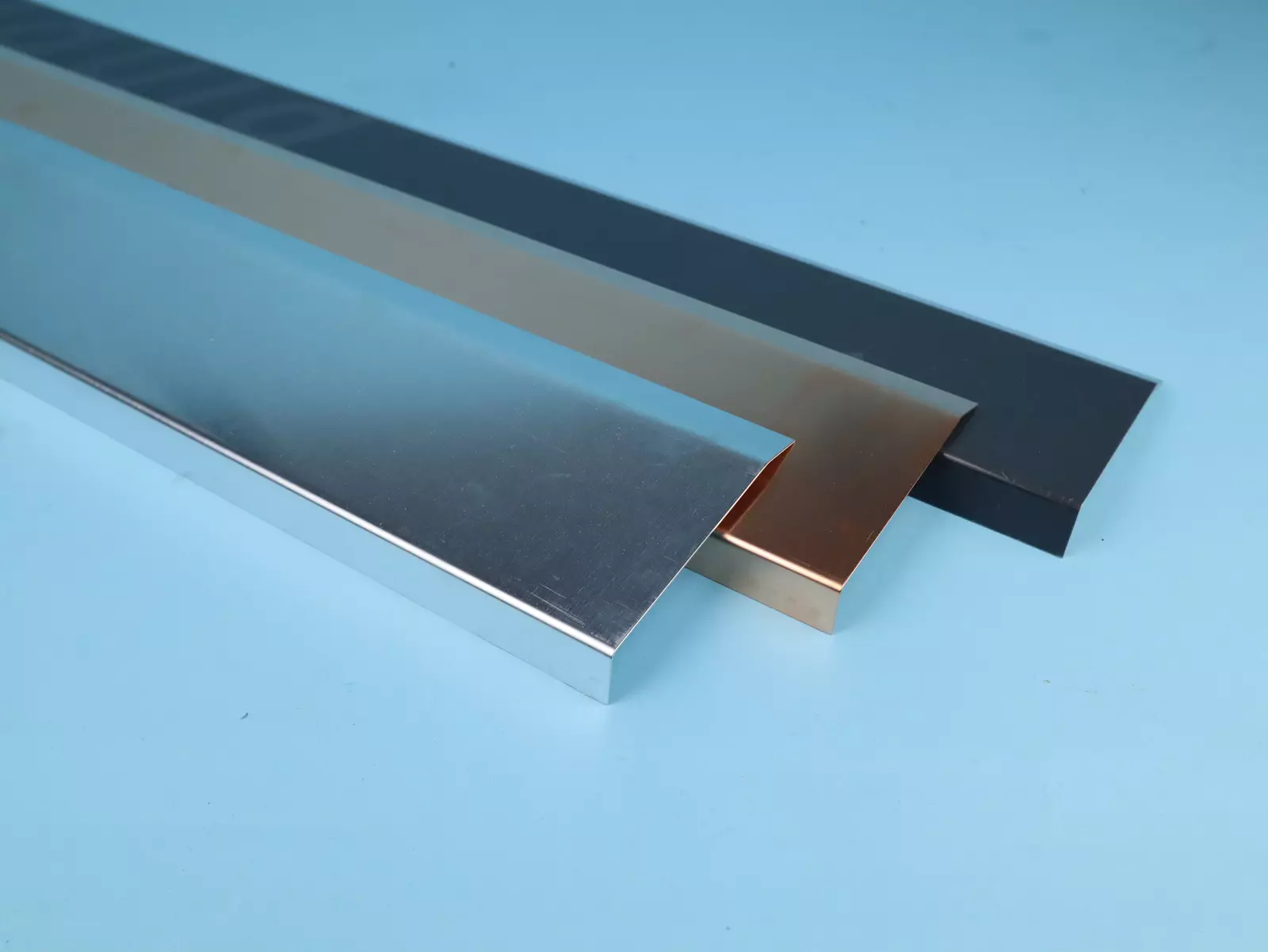 Drei Metallprofile in unterschiedlichen Farben liegen nebeneinander auf einer blauen Oberfläche. Die Profile sind silbern, kupferfarben und schwarz, mit glatten Oberflächen und rechteckigen Querschnitten.