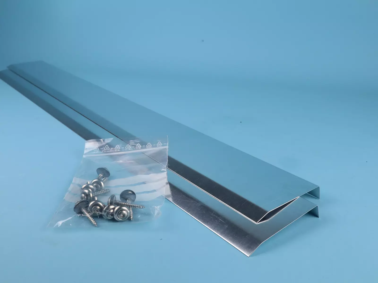 Zwei glänzende, metallene Profilbleche liegen parallel auf einem hellblauen Untergrund, neben einer Tüte mit silbernen Schrauben und Unterlegscheiben, die für die Montage benötigt werden.