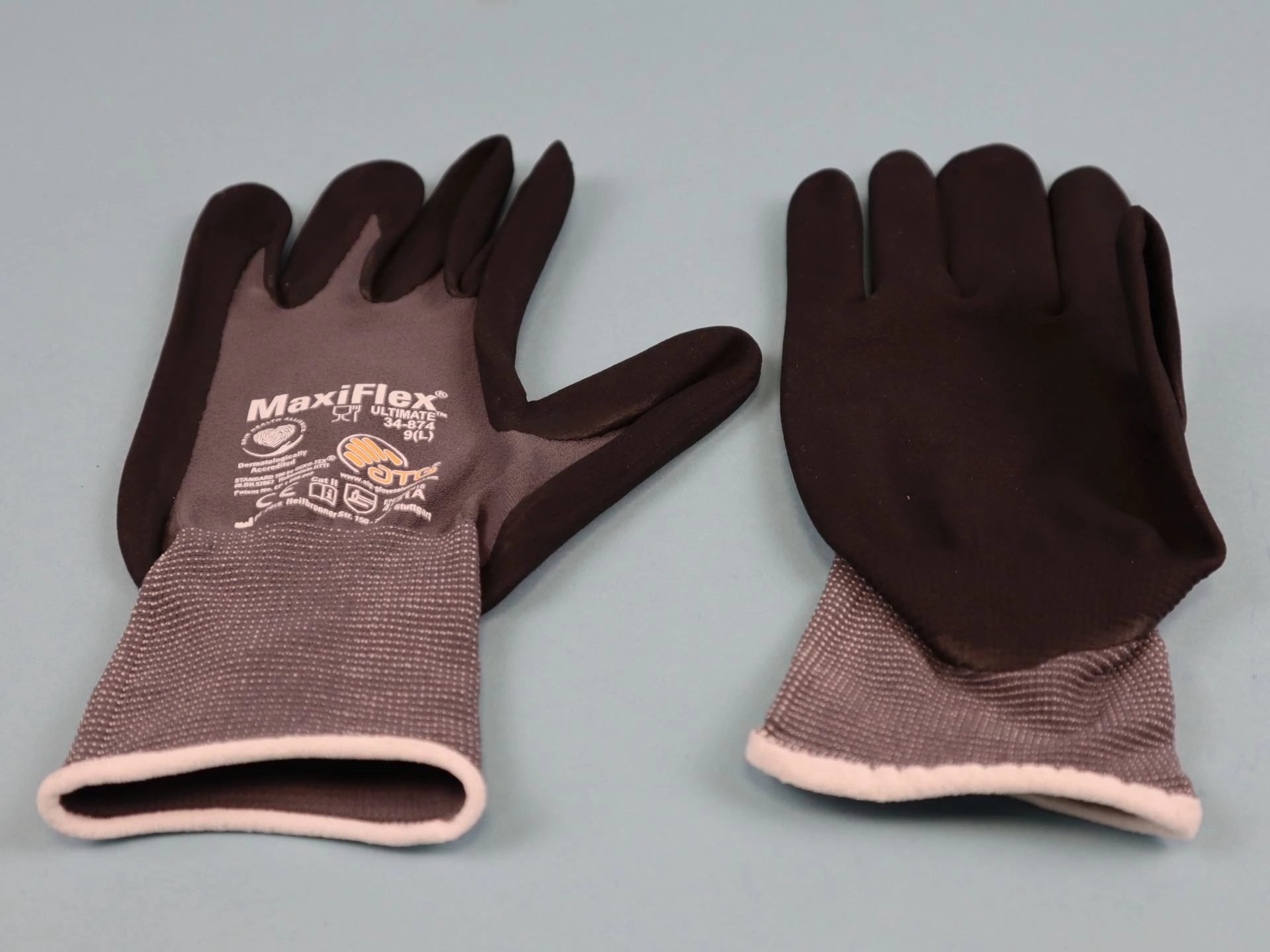 Paar graue MaxiFlex Ultimate Arbeitshandschuhe mit grauem Strickbund und rosa Innenfutter, palm- und fingerspitzenbeschichtet mit Nitril, hoher Tragekomfort, EN388-zertifiziert für Bau, Montage und Feinarbeiten.