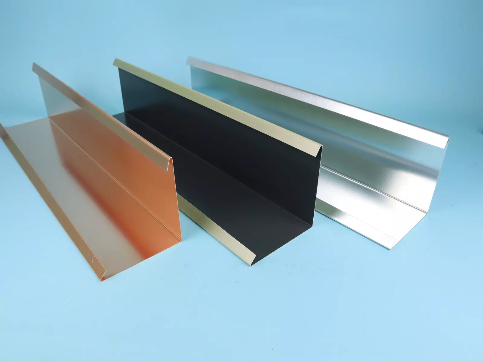 Drei unterschiedliche Metallprofile, in Kupfer, Schwarz und Silber, liegen nebeneinander auf einer hellblauen Hintergrundfläche. Die Profile haben eine ausgeprägte Form mit hohen Kanten und sind für Dachkonstruktionen geeignet.