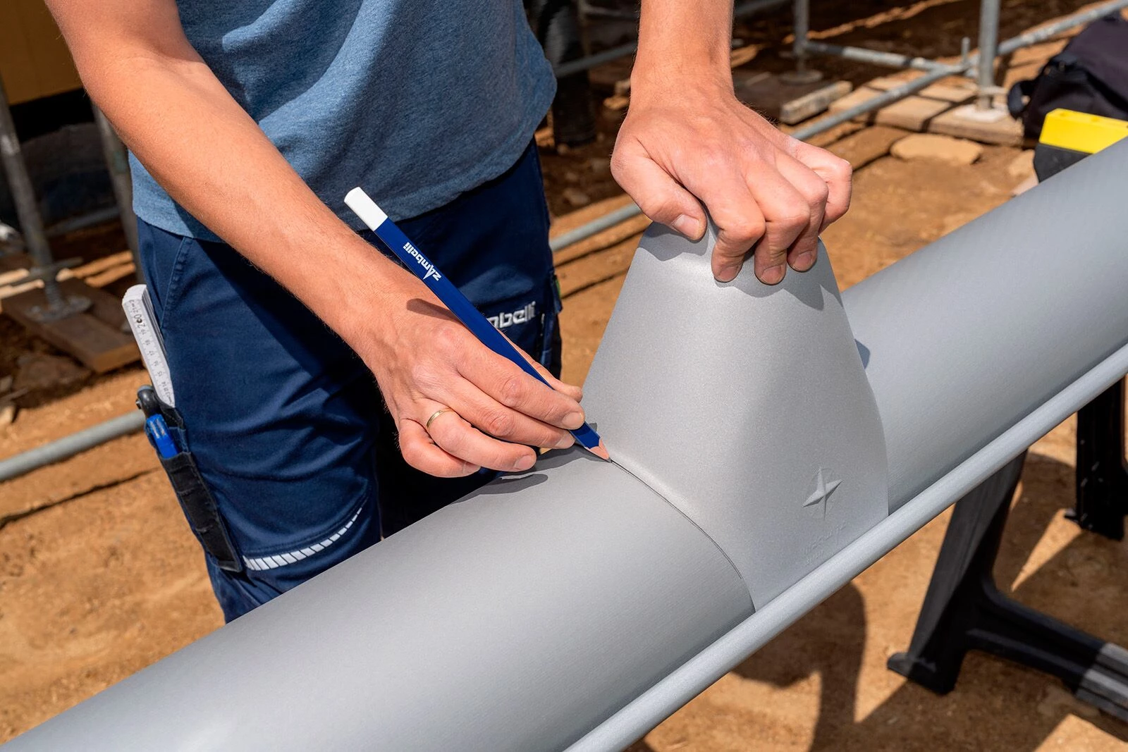 Eine Person zeichnet mit einem blauen Bleistift auf einer grauen Dachrinne, während sie die Rinne mit einer Hand festhält. Im Hintergrund sind Baustellenmaterialien und ein Erdgeschoss sichtbar.