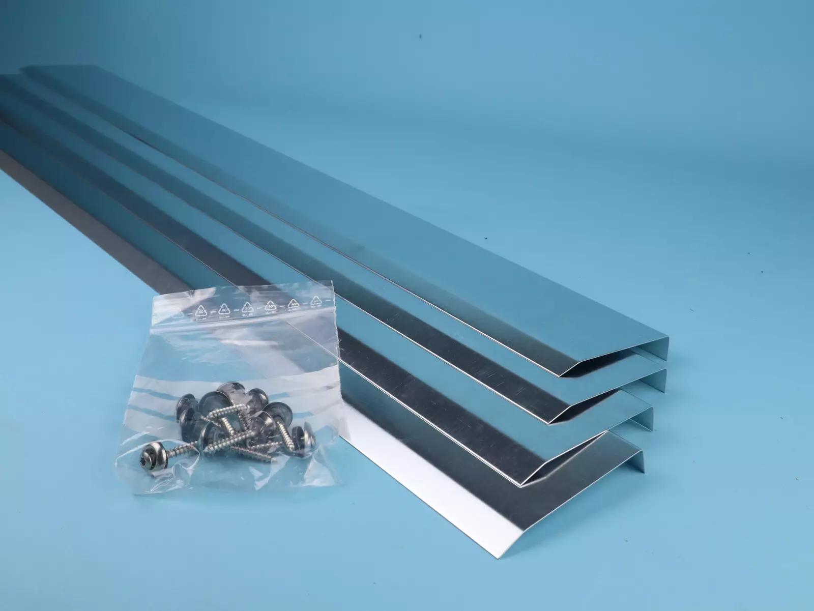 Vier glänzende Metallprofile mit rauen Kanten liegen parallel auf einem blauen Untergrund. Neben ihnen befindet sich ein transparenter Beutel mit mehreren Schrauben.