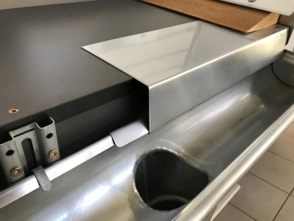 Ein silberner Dachrinnenabschnitt zeigt eine Metallplatte, die schräg aufliegt und teilweise eingeklemmt ist. Im Hintergrund ist eine Holzplatte sichtbar, während die Rinne auf einem fliesenförmigen Boden positioniert ist.