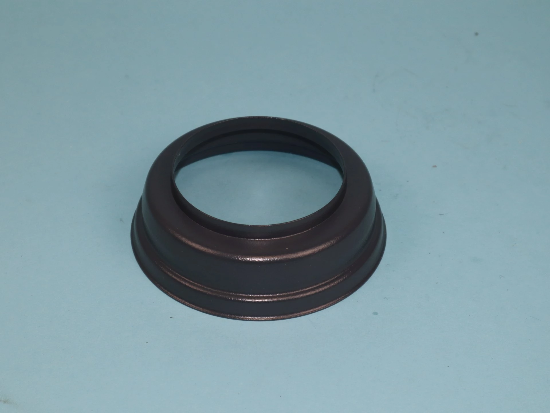 Ein schwarzer Metallring mit einer erhöhten Kante und einem zentralen Loch. Der Ring hat eine glatte Oberfläche und steht auf einer blauen Unterlage, ohne weitere Objekte im Umfeld.