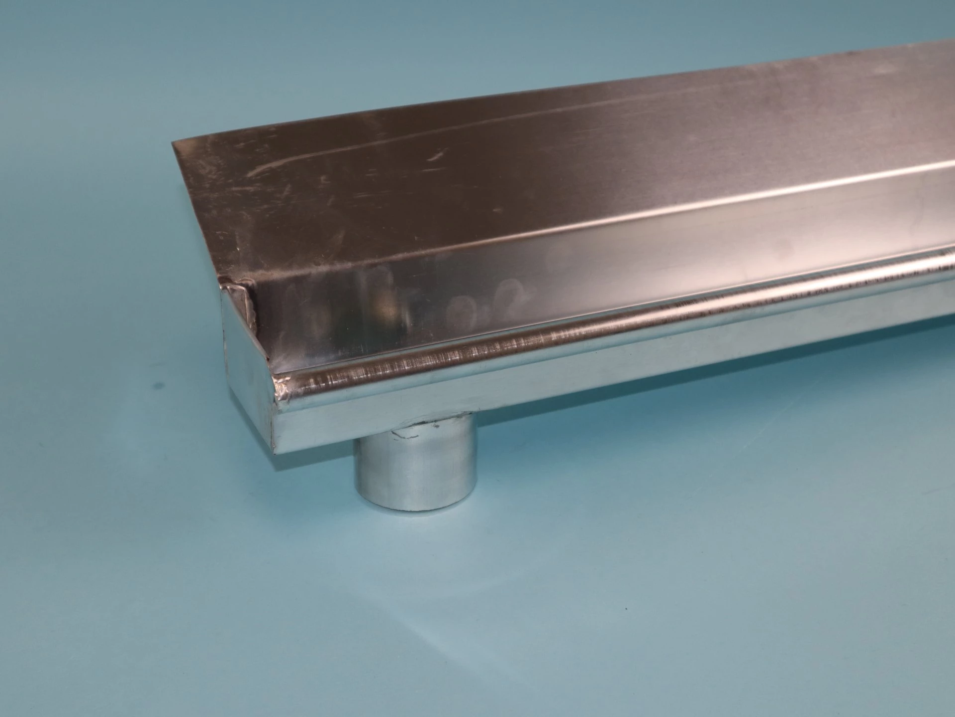 Ein glänzendes, rechteckiges Metallprofil aus Aluminium mit einer abgerundeten Kante und einem seitlichen Rohransatz. Es liegt auf einer hellblauen Unterlage.