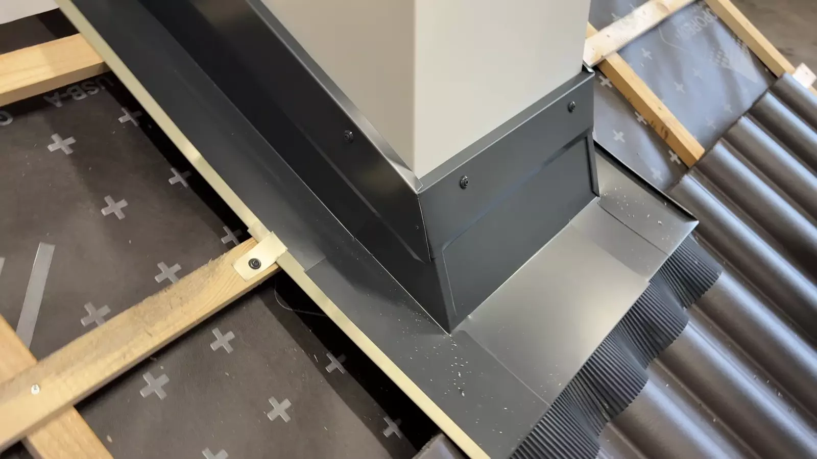 Ein Dachabdeckungsdetail zeigt eine graue Metallverkleidung um einen Schornstein, montiert auf einem geneigten Dach mit Wellblech. Holzlatten unterstützen die Struktur, während der Untergrund eine Schicht Schalung oder Geotextil aufweist.