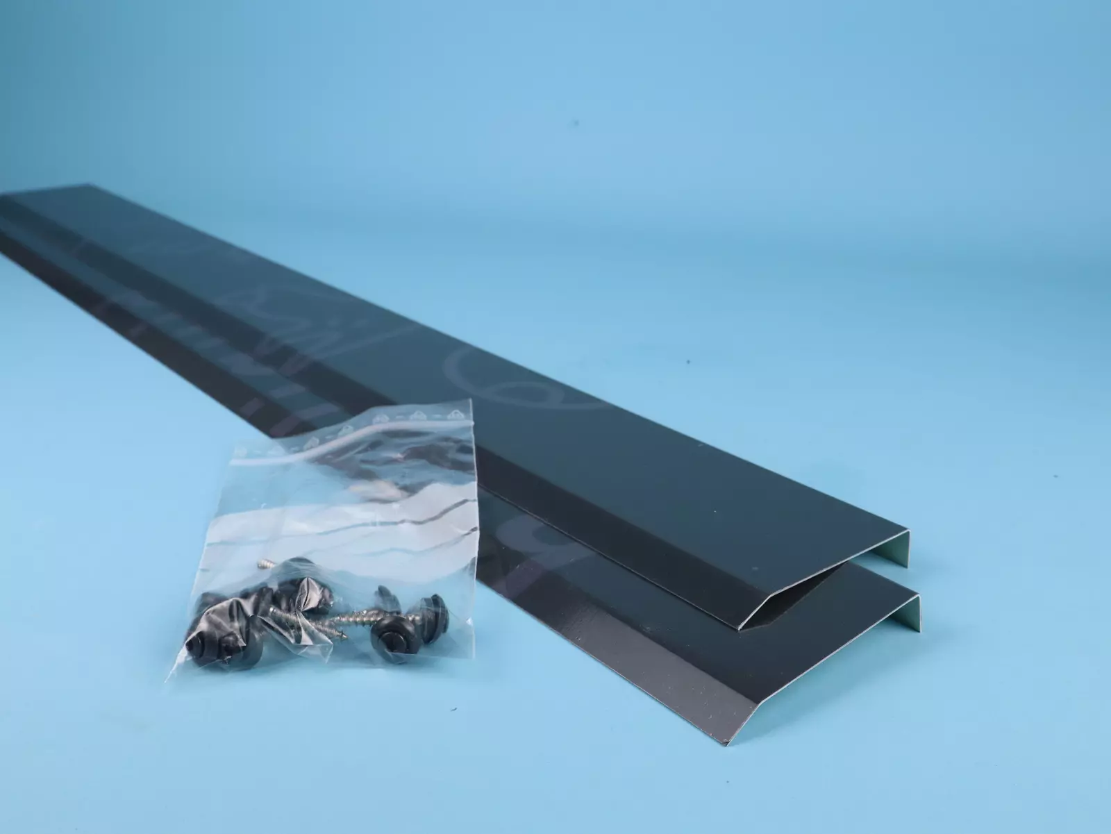 Zwei längliche Metallprofile mit einem gebogenen Ende liegen auf einer hellblauen Oberfläche. Daneben befindet sich ein durchsichtiger Beutel mit mehreren schwarzen Schrauben und Unterlegscheiben.