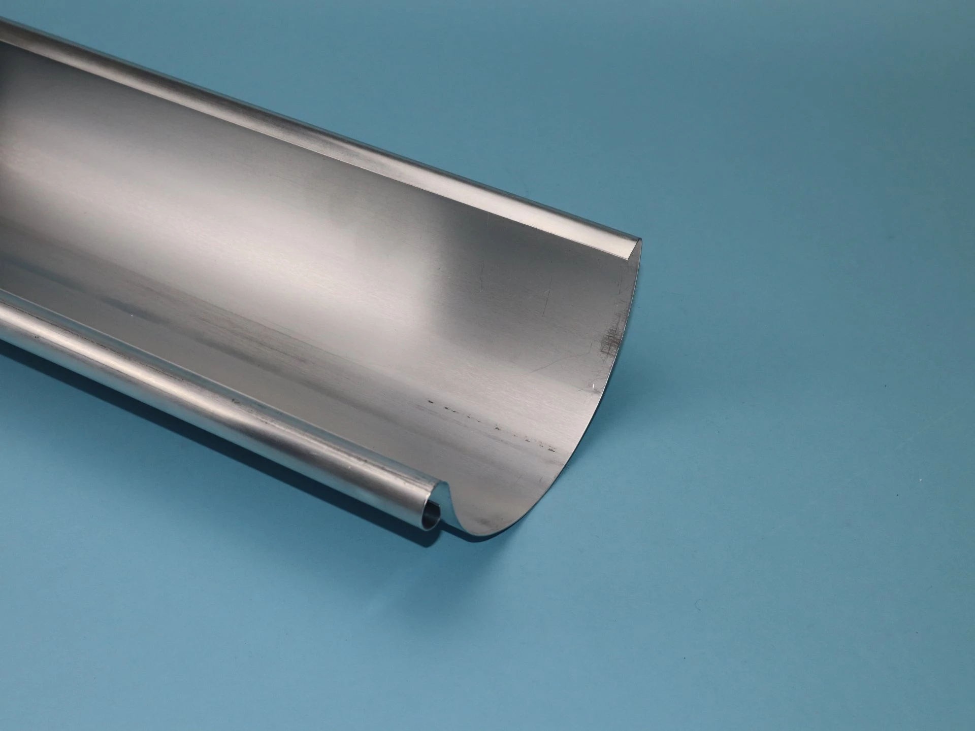 Ein längliches, glänzendes Metallblech mit geschwungener Kante liegt auf einem blauen Hintergrund. Es weist eine glatte Oberfläche und eine gebogene Rückseite auf, die für den Einsatz in Dachentwässerungssystemen geeignet ist.