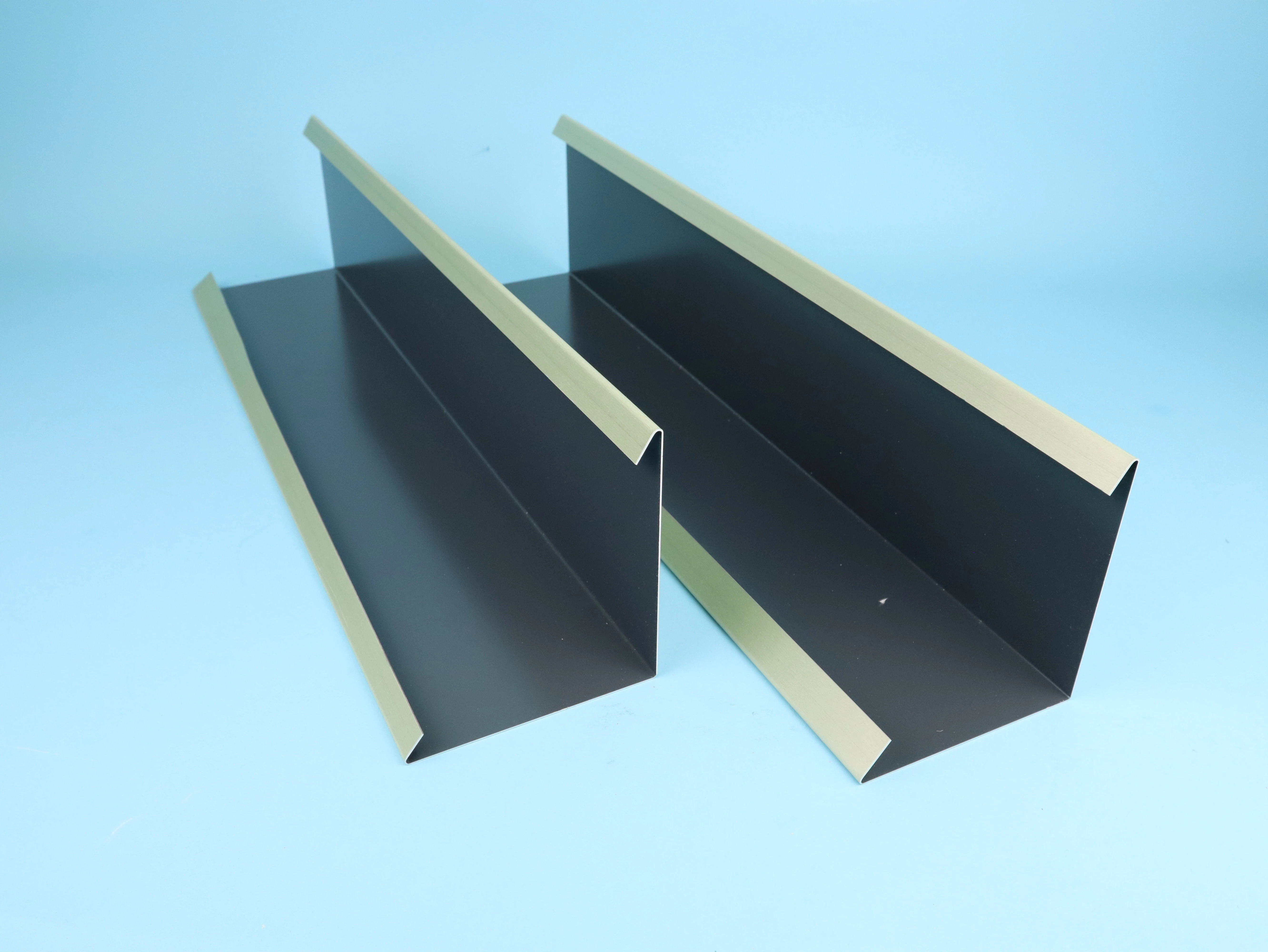 Zwei glänzende Aluminiumprofile in Schwarz mit goldenen Kanten liegen nebeneinander. Sie sind flach und länglich, geeignet für architektonische Anwendungen oder als Teil von Dachentwässerungssystemen.